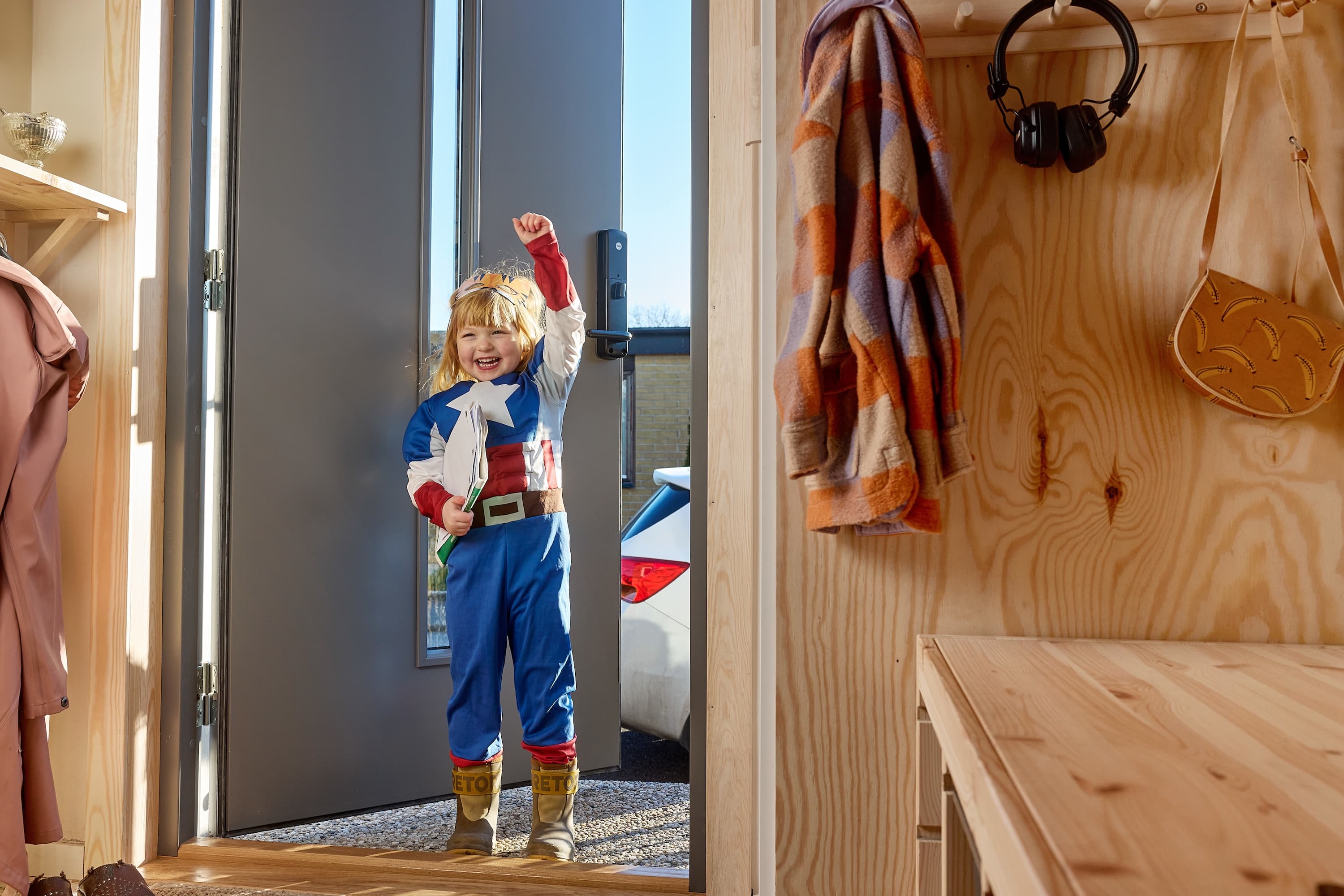 BoKlok Child in a superhero costume excitedly entering a room with wooden shelves holding clothes, a bag, and headphones.